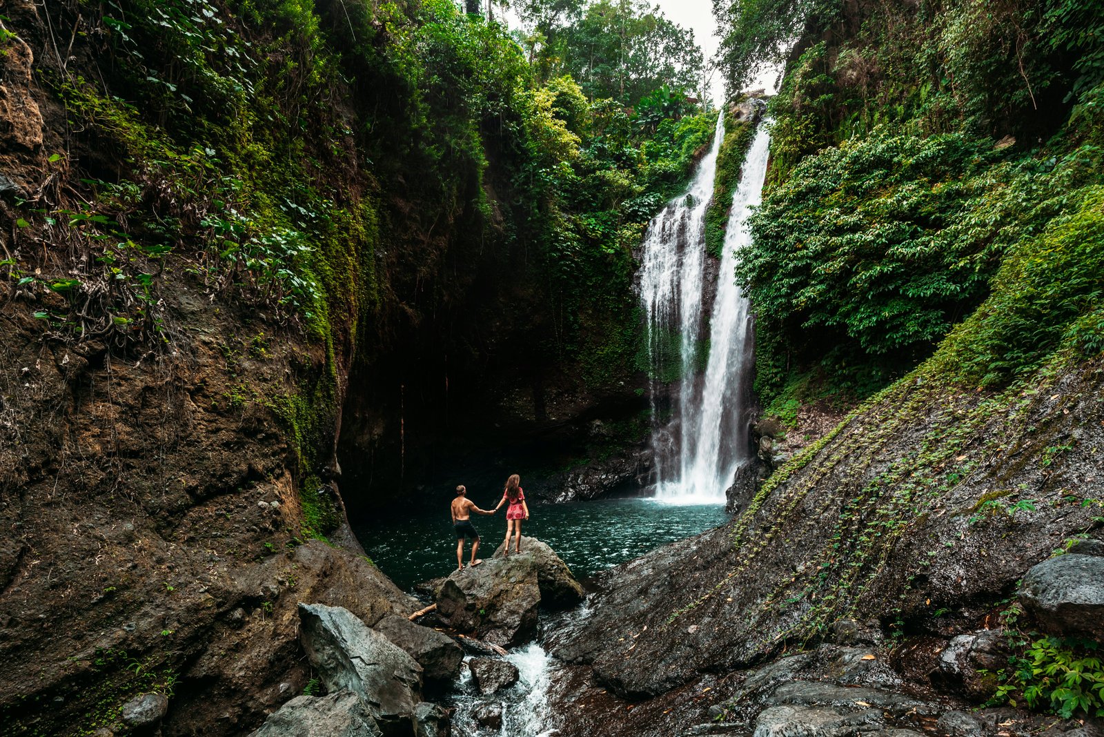 Un large coup d'un couple se tenant la main alors qu'ils regardent une grande cascade versant dans une piscine entourée de rochers de la jungle verte au-dessus